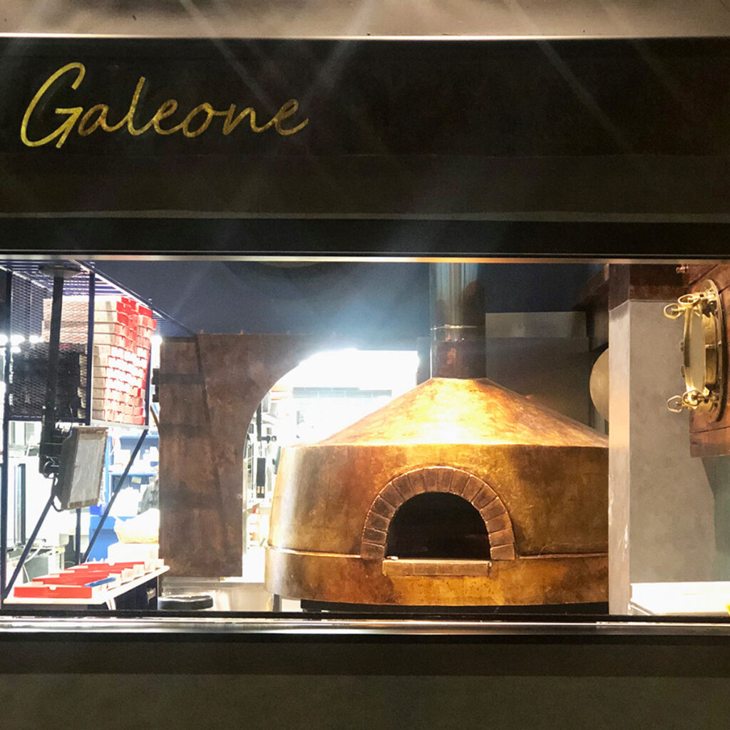 Παραδοσιακός ξυλόφουρνος στο εστιατόριο Galeone, με χάλκινη καμπύλη κατασκευή και άνοιγμα από τούβλα, μέσα σε μοντέρνο χώρο κουζίνας.